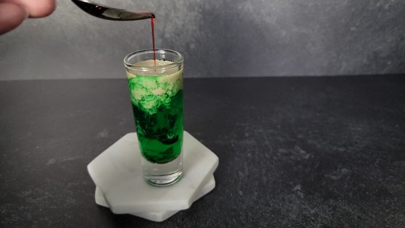 Spoon above a shot glass with red liquid dripping off of it into a green and cream liquid shot