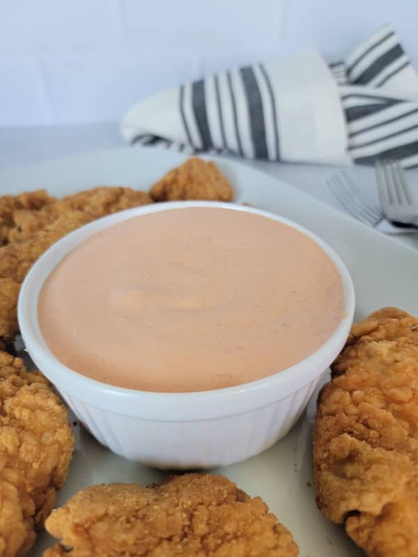 Mayo substance in a white bowl next to chicken tenders on a white plate and a cloth napkin