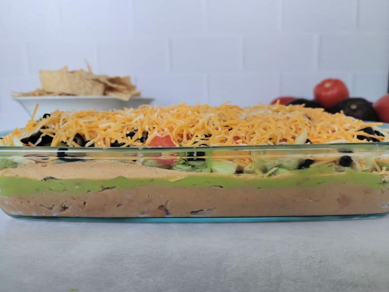 Layered dip in a glass baking dish with shredded cheese on top next to a bowl of tortilla chips