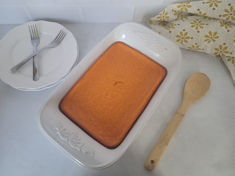 baked carrot souffle in a casserole dish next to a wooden spoon and plate with forks