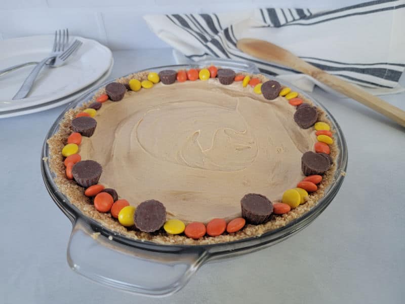 peanut butter pie rimmed with Reese's pieces in a glass baking dish on a counter with plates and forks in the background next to a cloth napkin