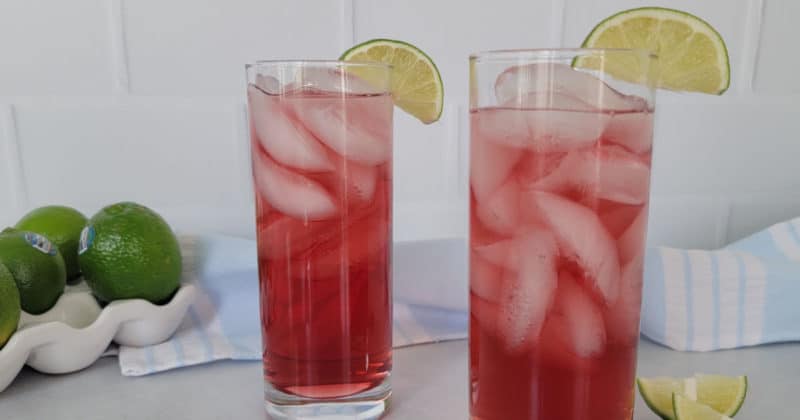 Two red woo woo drinks in tall glasses with lime wedges next to a stack of limes