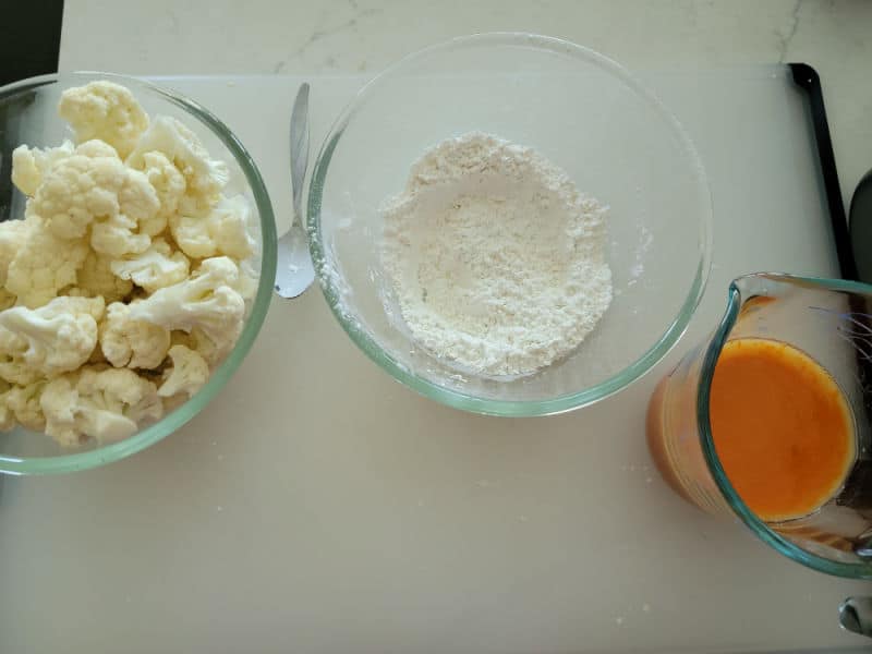 cauliflower florets in a glass bowl next to a bowl of flour and a bowl of buffalo sauce