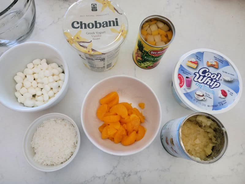 mini marshmallows, coconut, greek yogurt, fruit in a can, cool whip, pineapple, and mandarin oranges in a white bowl on a counter