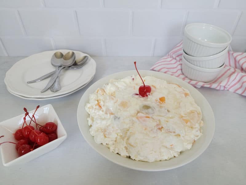 Ambrosia fruit salad in a white bowl next to a bowl of maraschino cherries, a stack of plates with forks, and a stack of small bowls on a cloth napkin