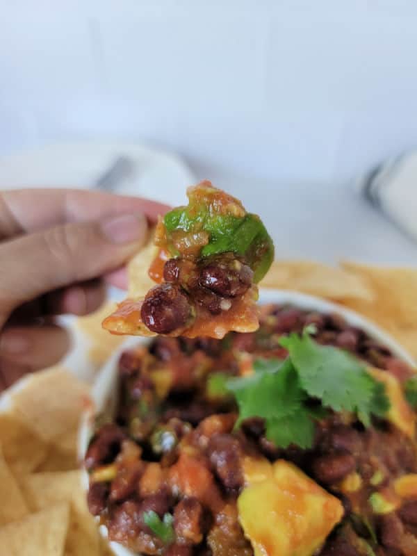 hand holding a tortilla chip with black bean salsa on it over a white bowl with salsa surrounded by more chips