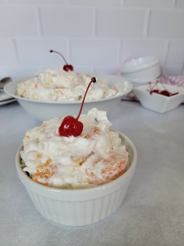 Small white bowl with ambrosia salad with a maraschino cherry on top in front of a large bowl of salad and a bowl of cherries