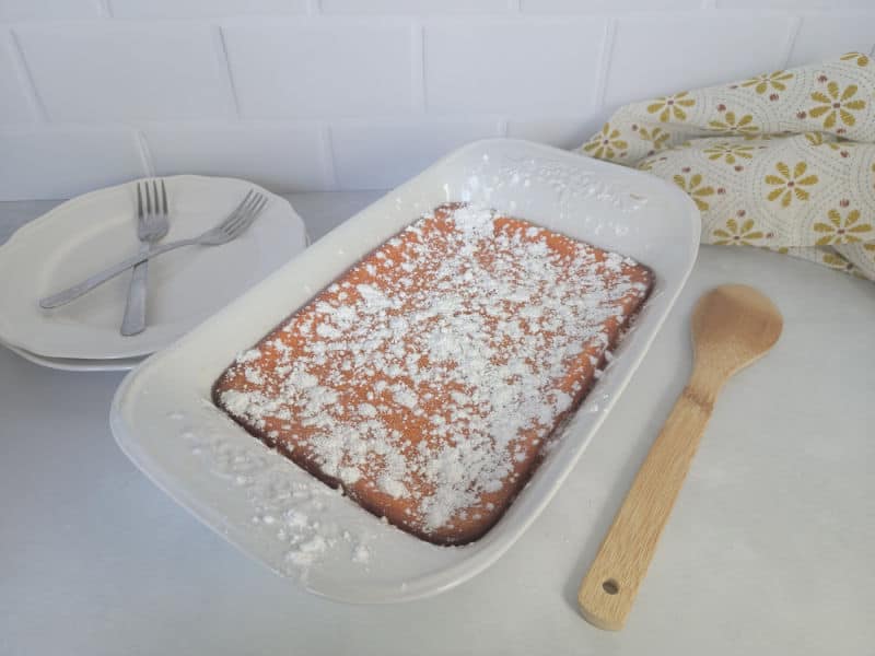 powdered sugar garnished carrot souffle in a casserole dish with a wooden spoon next to plates and forks