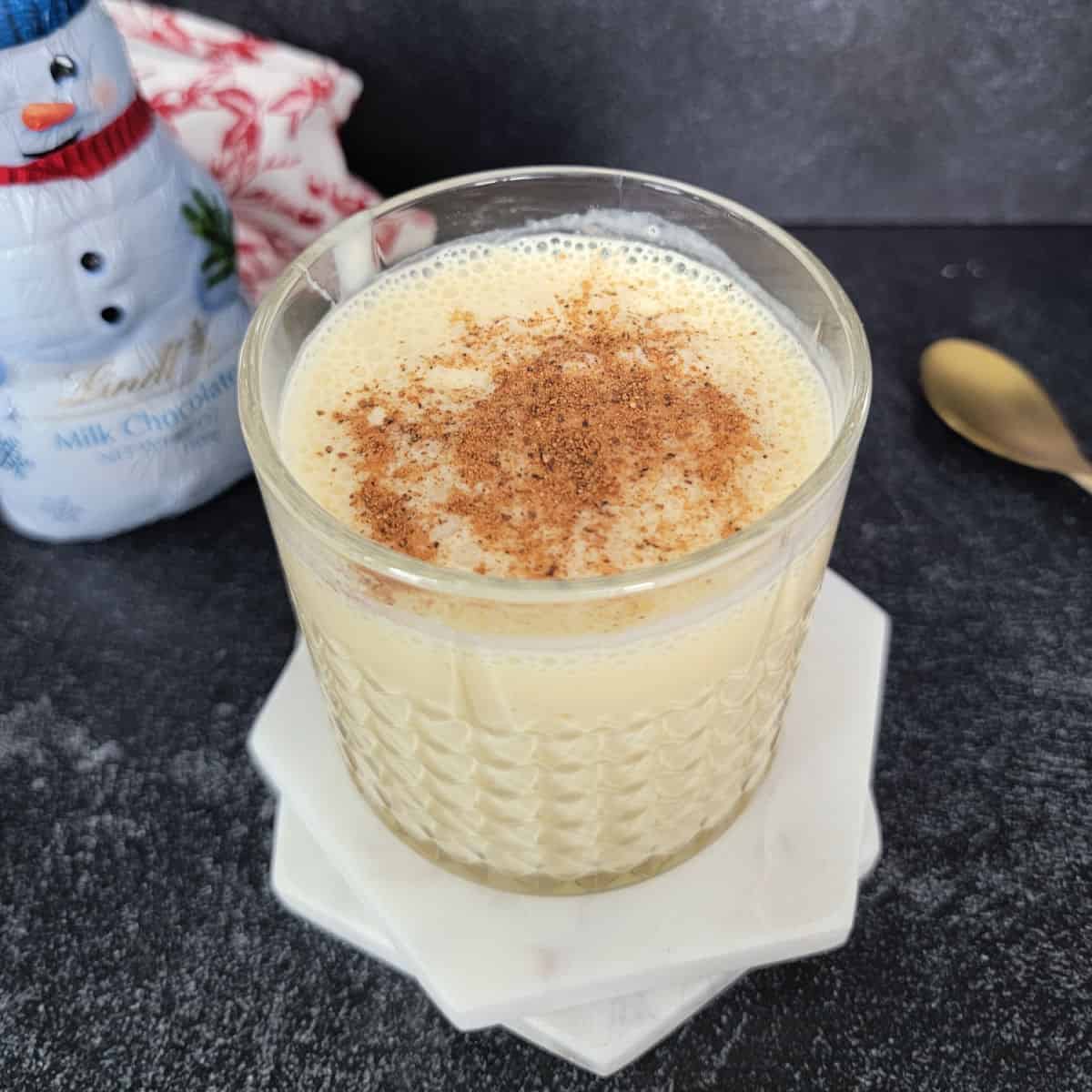 Bourbon Eggnog in a crystal glass on white coasters next to a gold spoon and snowman