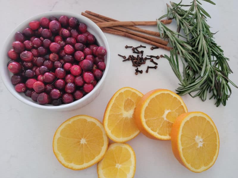 white bowl filled with cranberries, cloves, rosemary, cinnamon sticks, and orange wheels on a white counter for simmering Christmas potpourri