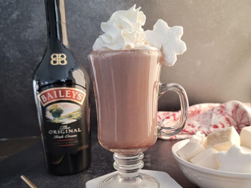 Bailey's Irish Cream Hot Chocolate in a glass mug with whipped cream and a star marshmallow next to a bottle of Baileys Irish cream