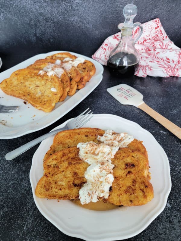 Eggnog French Toast slices on a white plate garnished with eggnog whipped cream and cinnamon/nutmeg next to a platter and a spatula