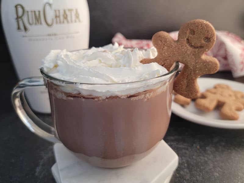 Glass mug with RumChata Hot Chocolate garnished with whipped cream and a gingerbread cookie with a bottle of RumChata in the background