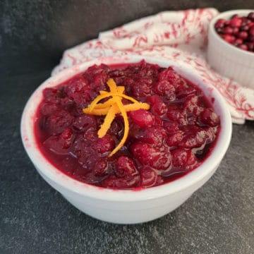 homemade cranberry salsa in a white bowl with an orange zest garnish next to a bowl of cranberries and a red cloth towel