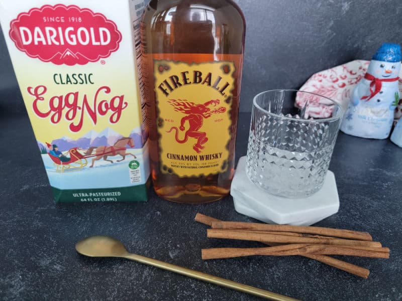 Classic eggnog, fireball bottle, glass, cinnamon sticks, and a gold spoon