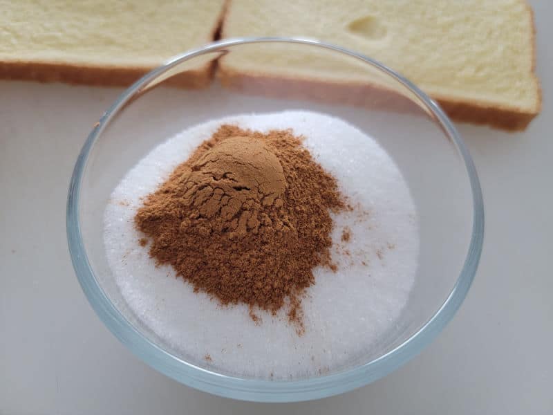 Cinnamon on top of sugar in a glass bowl near bread. 