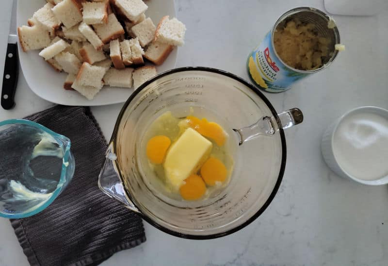 butter and eggs in a glass mixing bowl next to cut up bread pieces, and pineapple in a can