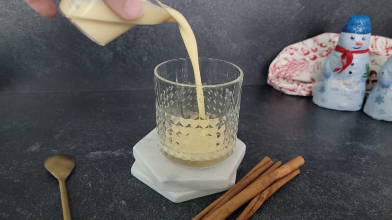 eggnog pouring into a glass next to cinnamon sticks and a spoon
