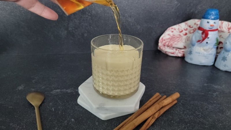 Fireball pouring into eggnog next to a spoon and cinnamon sticks. 