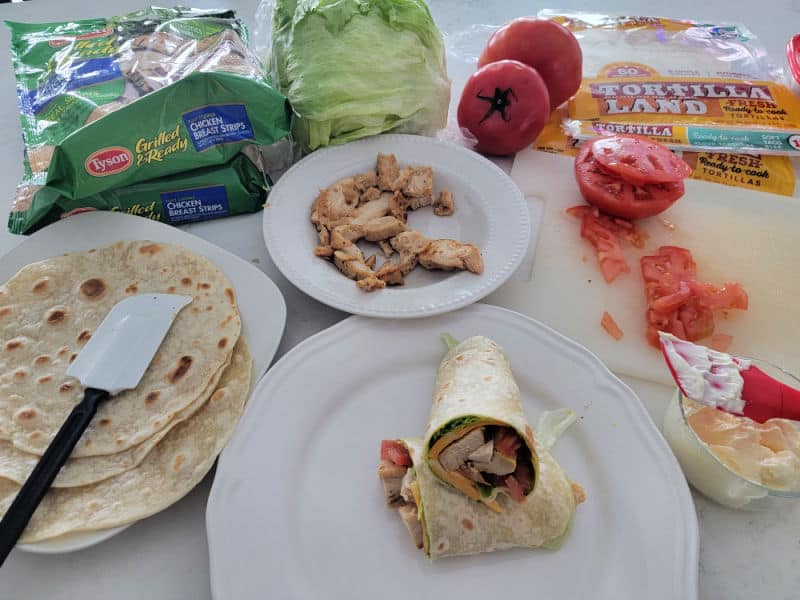 Chicken wrap on a white plate next to chicken, tortillas, diced tomatoes, and mayo