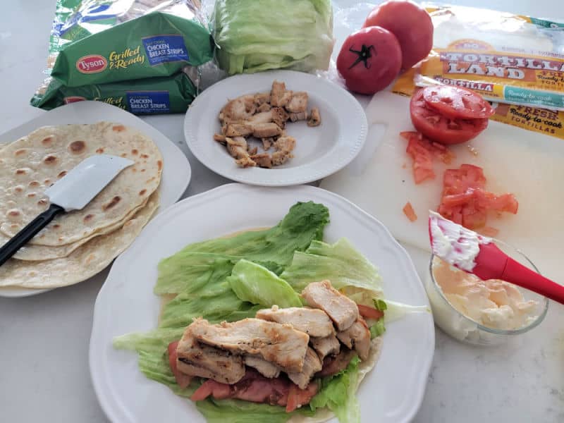 Chicken wrap on a white plate next to a plate of tortillas, chicken, tomatoes, and mayo