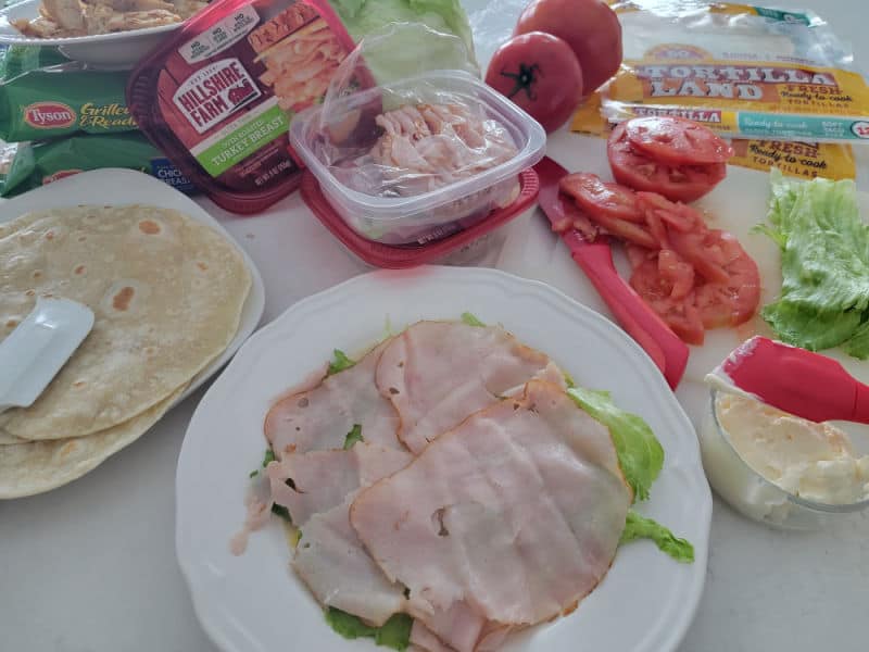 ham on lettuce and a tortilla next to a plate of tortillas, lettuce, tomatoes, mayo, and a knife