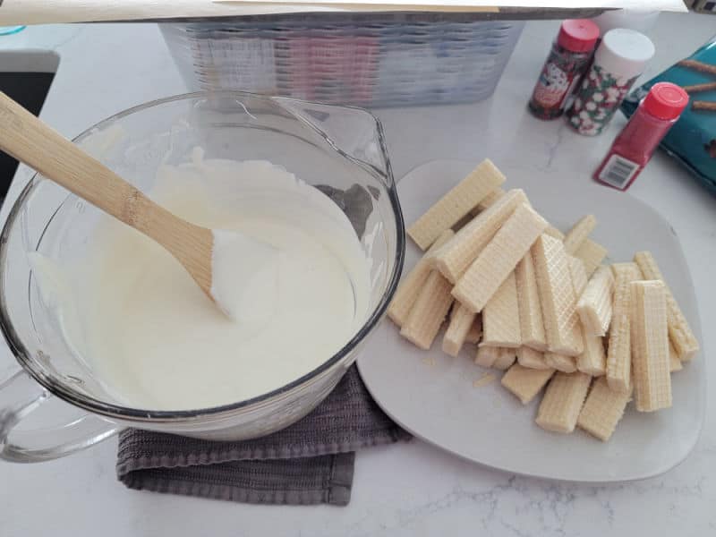 Melted vanilla almond bark in a glass bowl next to a plate of vanilla wafers and holiday sprinkles