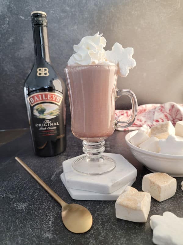 Baileys Hot Chocolate in a glass mug with whipped cream next to a bottle of Original Baileys Irish Cream, a bowl of marshmallows, a gold spoon, with a cloth napkin