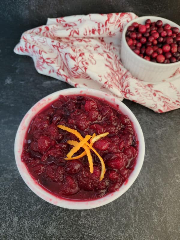 Homemade cranberry sauce in a white bowl with orange strips next to a bowl of fresh cranberries
