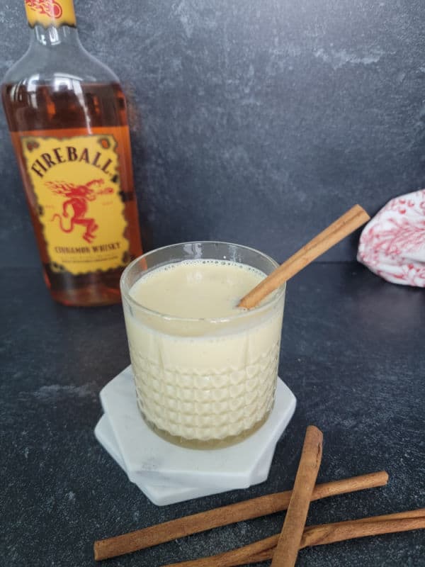 Fireball behind a glass of eggnog on a white coaster next to cinnamon sticks. 