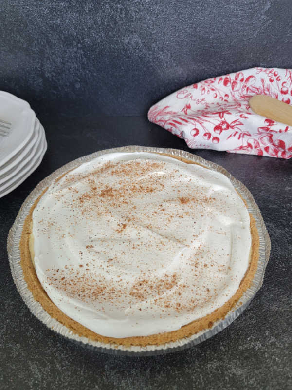 No bake eggnog pie in a graham cracker crust on a dark counter top next to plates and a cloth napkin