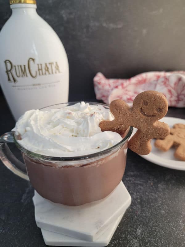 Glass mug with RumChata Hot Chocolate garnished with whipped cream and a gingerbread cookie with a bottle of RumChata in the background
