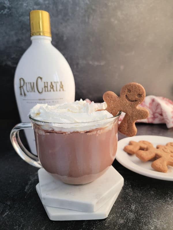 Glass mug with RumChata Hot Chocolate garnished with whipped cream and a gingerbread cookie with a bottle of RumChata in the background