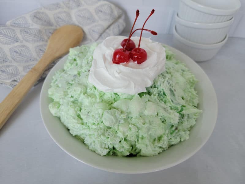 Classic Watergate Salad in a white bowl with a dollop of Cool Whip and Maraschino Cherries next to a wooden spoon and three white bowls