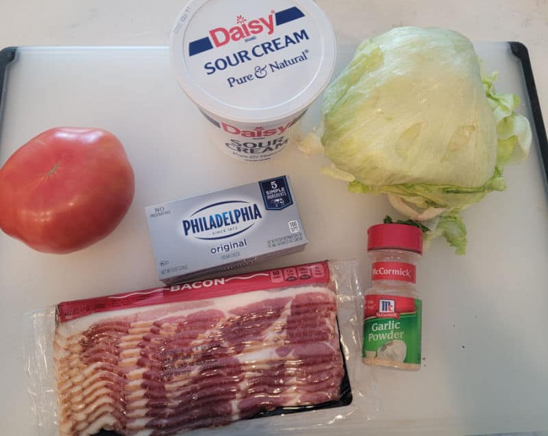 Sour cream, a head of lettuce, tomato, cream cheese, package of bacon, and garlic powder on a cutting board 
