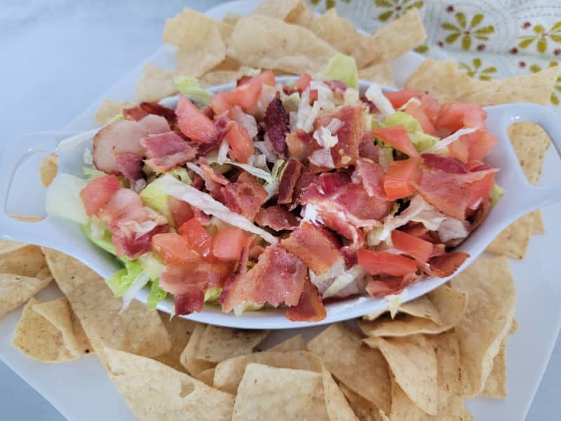 Diced bacon, diced tomatoes, and lettuce in a white bowl next to tortilla chips and a cloth napkin