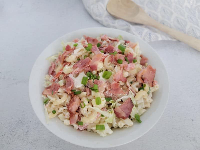 BLT Macaroni Salad in a white bowl next to a wooden spoon and cloth napkin 