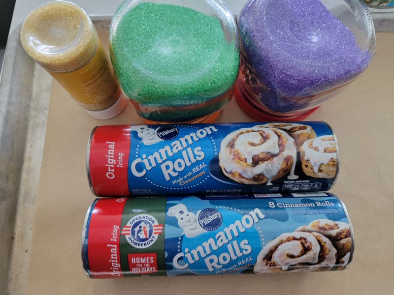 Pillsbury Cinnamon Roll packages next to gold, green, and purple sprinkles, on a parchment lined baking sheet
