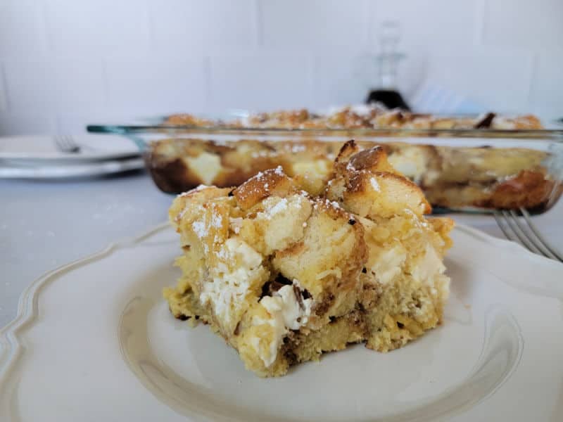 A piece of French Toast overnight breakfast casserole on a white plate with a full casserole dish behind it. 