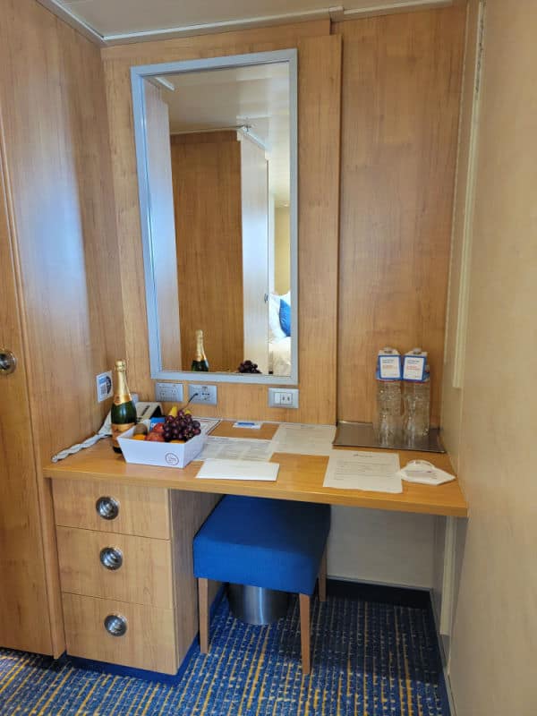 Built in desk with large mirror, bottle of champagne, fruit basket, and papers on the desk. 