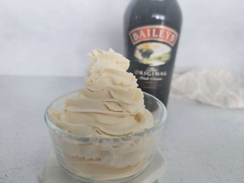 Baileys Whipped cream in a small glass bowl with a bottle of Baileys Irish Cream in the background next to a cloth napkin