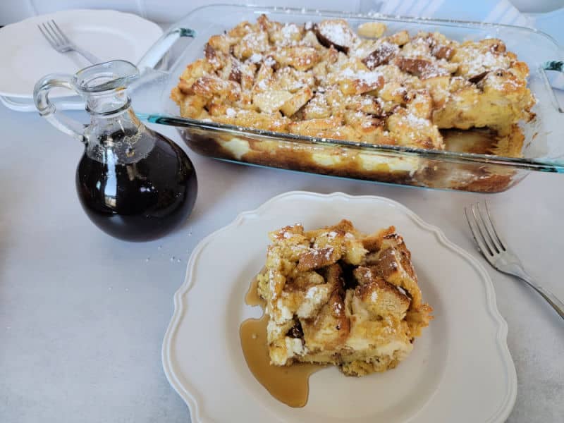 Overnight French Toast casserole in a baking dish next to maple syrup with one piece cut out on a white plate with a fork. 
