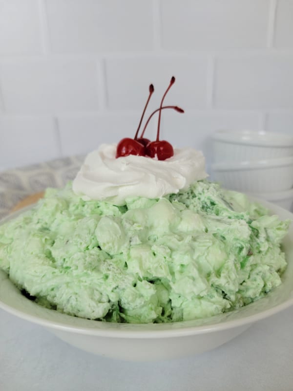 Classic Watergate Salad in a white bowl with a dollop of Cool Whip and Maraschino Cherries next to a wooden spoon and three white bowls