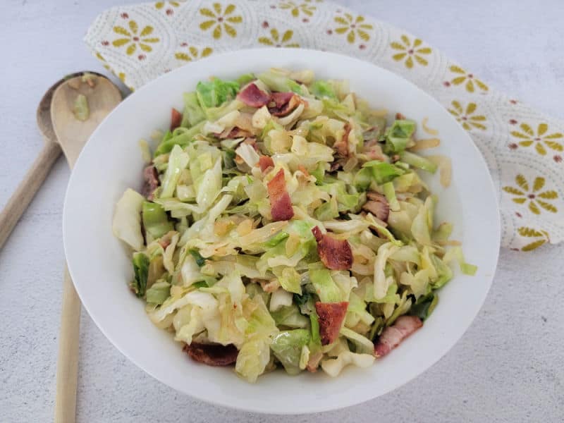 Fried cabbage with bacon in a white bowl next to wooden spoons and a cloth napkin 