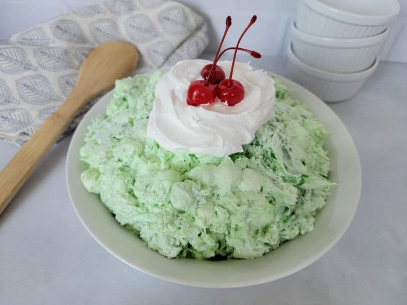 Easy Watergate Green Salad in a white bowl with a dollop of Cool Whip and Maraschino Cherries next to a wooden spoon and three white bowls