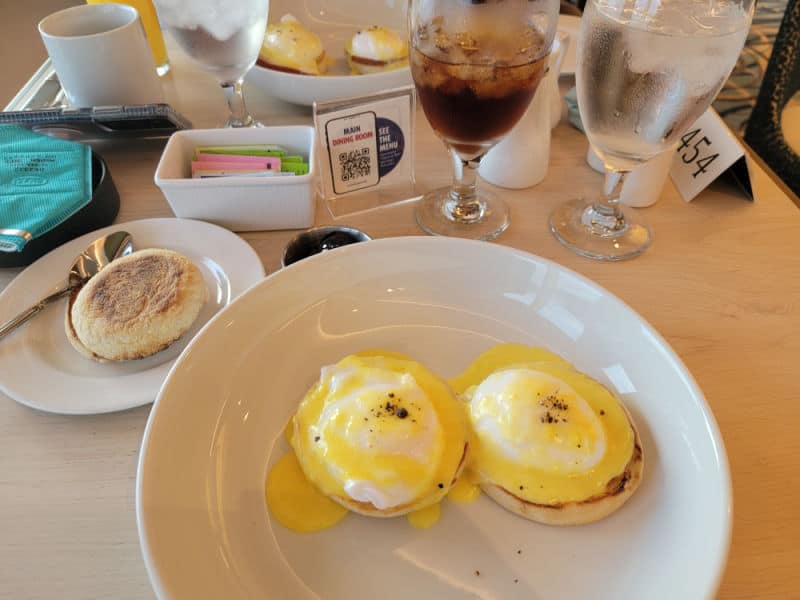 Eggs benedict on a white plate next to a English Muffin and glass of iced tea