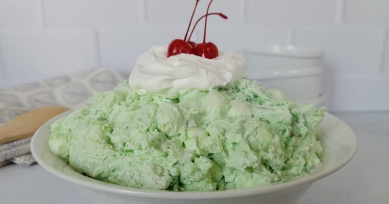 Classic Watergate Salad in a white bowl with a dollop of Cool Whip and Maraschino Cherries next to a wooden spoon and three white bowls