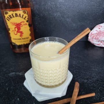 Fireball Eggnog in a crystal glass with cinnamon stick garnish in front of a bottle of Fireball Cinnamon Whiskey