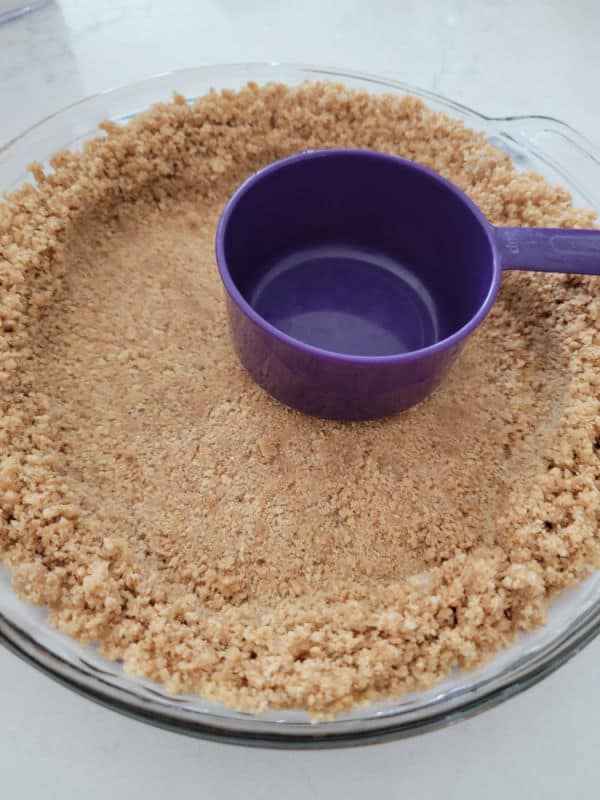 Graham Cracker Crust in a glass pie pan with a purple measuring cup pressing graham cracker crumb mixture