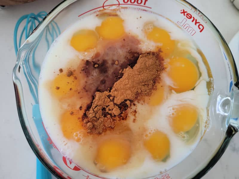 Large Pyrex measuring bowl with eggs, milk, and ground cinnamon next to a blue whisk for French Toast Overnight Cassserole